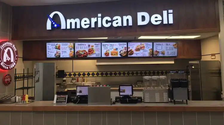 Interior of American Deli restaurant featuring an illuminated “American Deli” sign, digital menu boards with food items, visible kitchen space, and a counter with register screens and a drink dispenser.