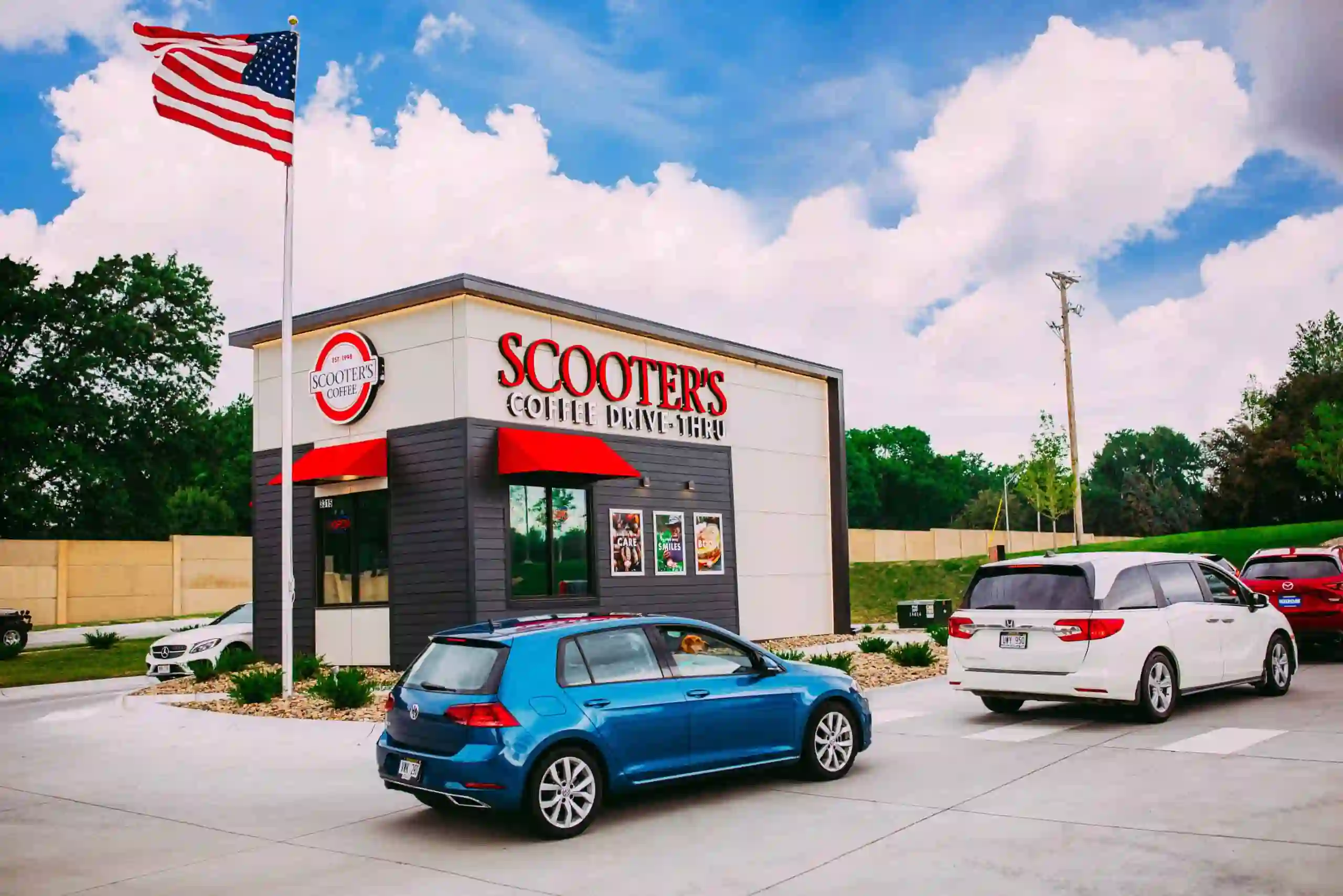 Scooter’s Coffee drive-thru location with red signage, white and dark gray exterior, American flag, and several cars in line under a partly cloudy sky.