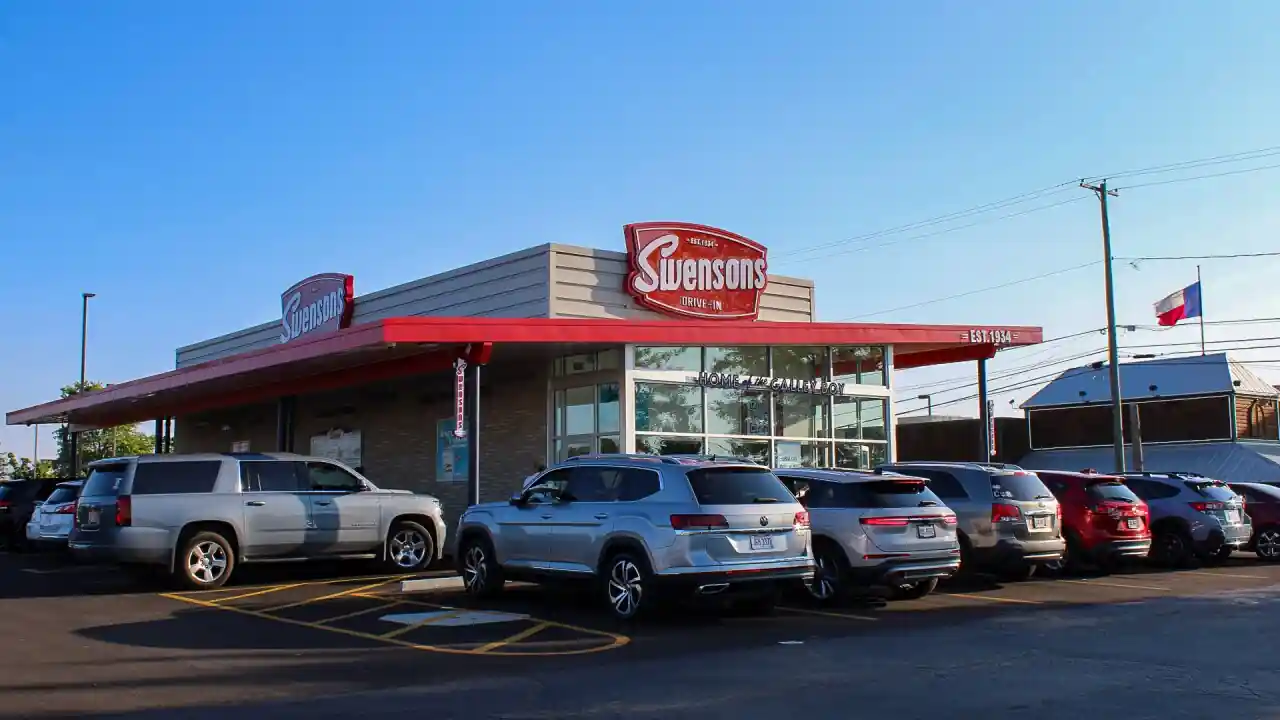 Front view of Swensons Drive-In restaurant with red signage on the roof, large windows, a red awning, and multiple parked cars in front under an overcast sky.