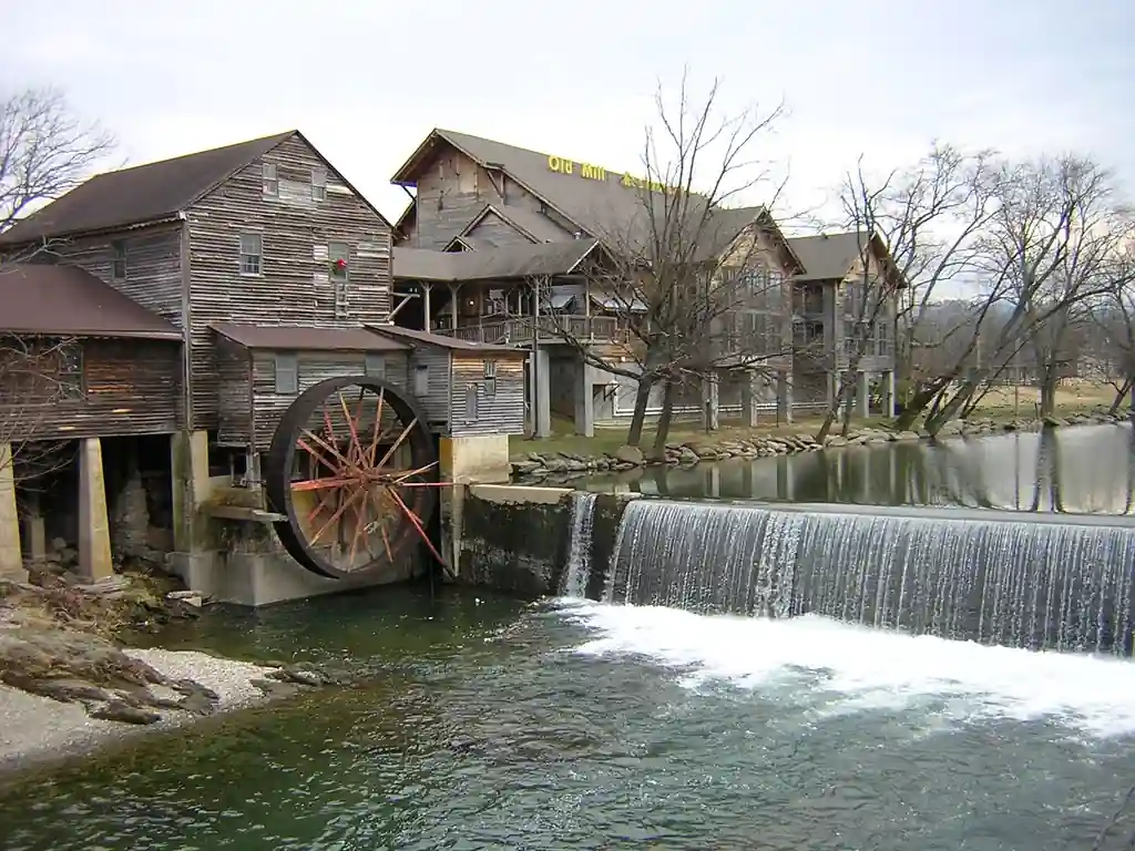 Historic wooden mill with a large water wheel beside a cascading waterfall, surrounded by bare trees and a grassy landscape.