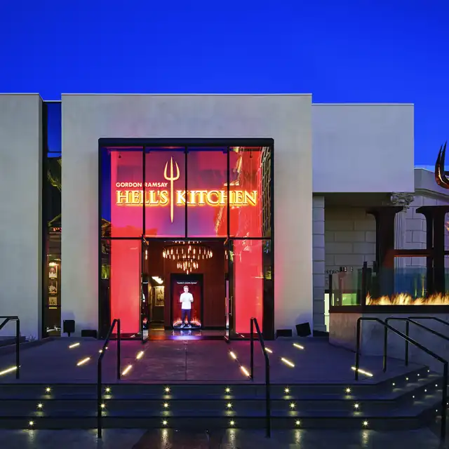 Exterior of Hell’s Kitchen restaurant with large glass entrance, glowing red backdrop, yellow “Hell’s Kitchen” sign, pitchfork symbol, and a chef statue inside the foyer—highlighting dramatic and upscale dining ambiance.