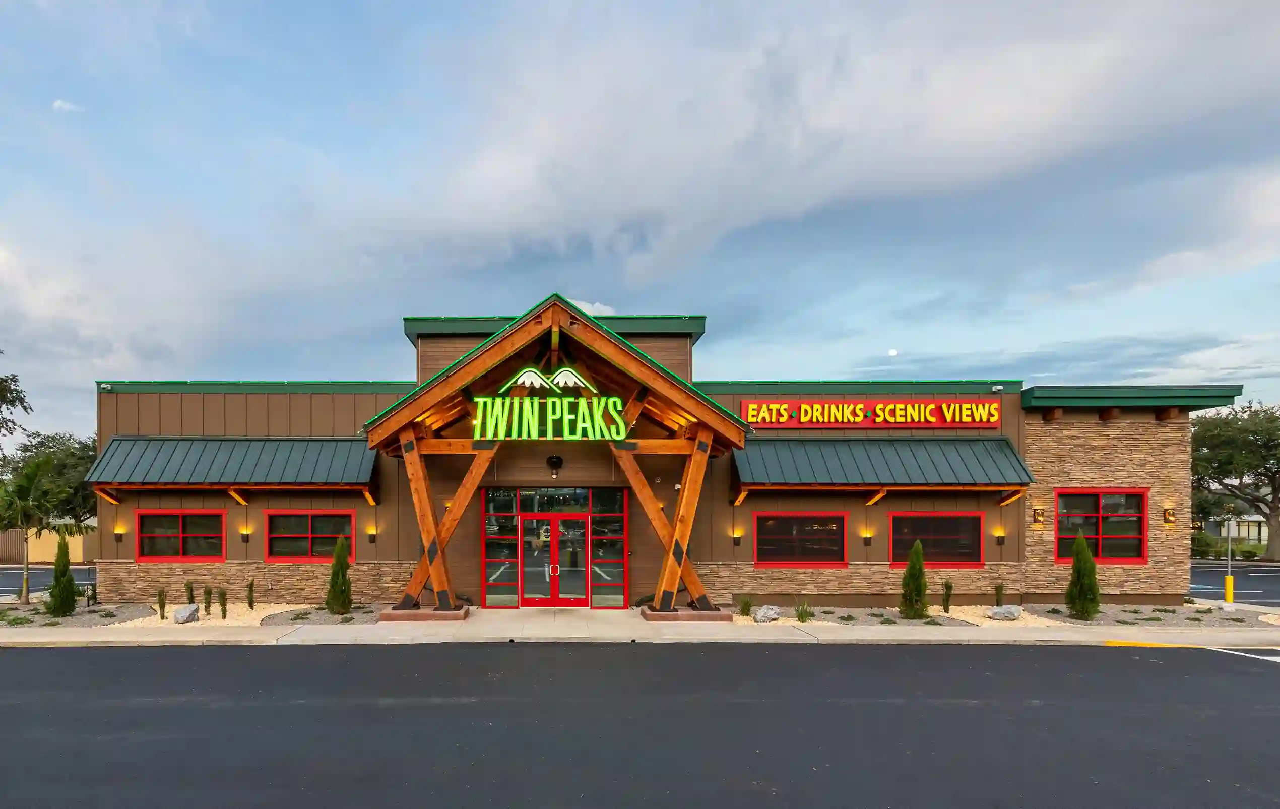Exterior of Twin Peaks restaurant showing green neon “TWIN PEAKS” sign, red-framed windows, stone and wood facade, green metal roof, and signage reading “EATS • DRINKS • SCENIC VIEWS” above the entrance, surrounded by a paved lot and landscaping.