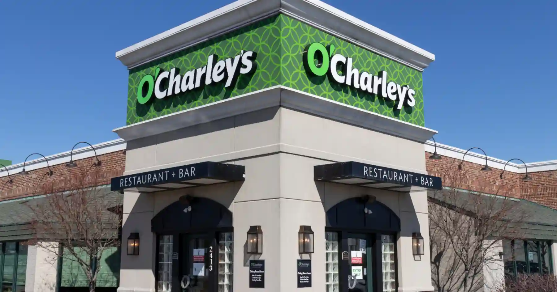 O’Charley’s Restaurant exterior with green circular signage, brick and beige walls, dual black awnings labeled “Restaurant + Bar,” and glass entrance marked with address number 2413.