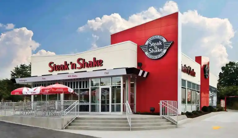 Exterior of Steak 'n Shake restaurant with red-and-white Coca-Cola umbrellas, glass entrance, bold red signage, and landscaped walkway on a clear day.