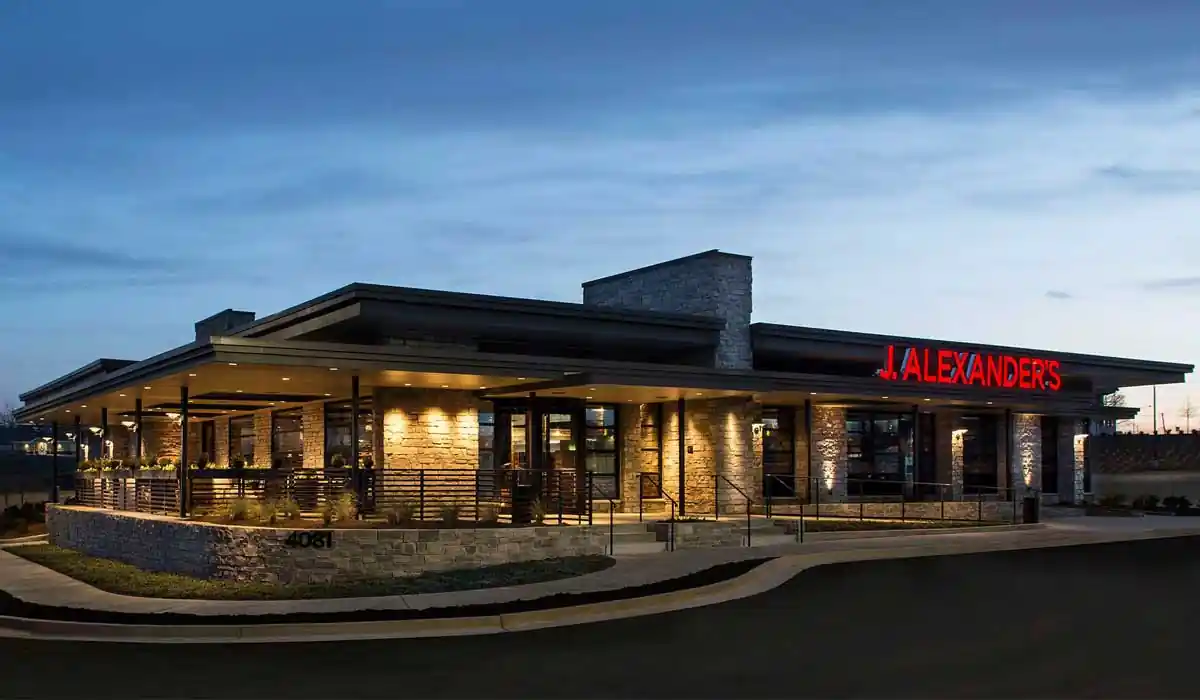 J. Alexander’s Restaurant exterior at dusk, with red illuminated signage, stone walls, large glass windows, and covered outdoor seating area lit warmly.