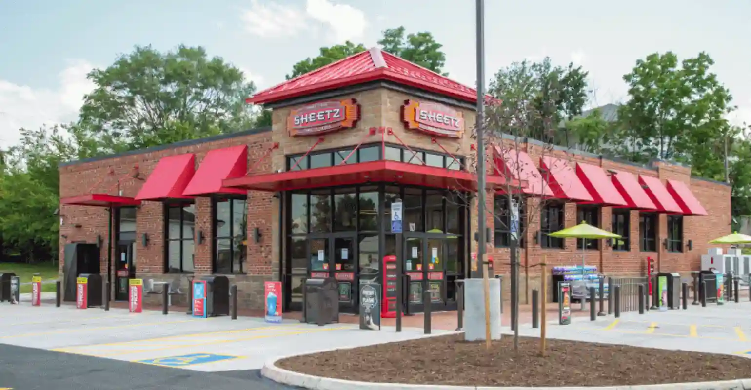 Exterior view of Sheetz convenience store and gas station showing red awnings, red signage above doors and windows, brick building design, patio seating with green umbrellas, landscaped bushes and trees, and a designated handicapped parking spot under clear skies.