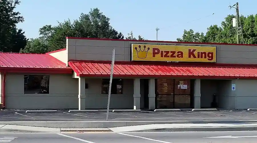Exterior of Pizza King restaurant with gray walls, red gable roof, yellow “Pizza King” sign featuring crown logo above entrance, two front windows, and street-side location with visible trees and sidewalk.