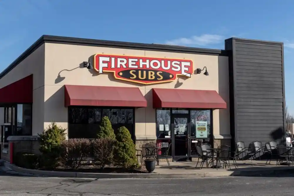 Daytime image of Firehouse Subs restaurant exterior showing red awnings, prominent red and yellow sign above entrance, beige building facade, patio seating area, and landscaped bushes under a clear blue sky.