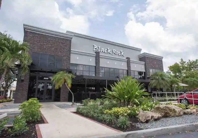 Modern exterior of Black Rock restaurant with dark brick walls, large glass windows, palm trees, and the number “8200” near the entrance; a red car is parked to the side.
