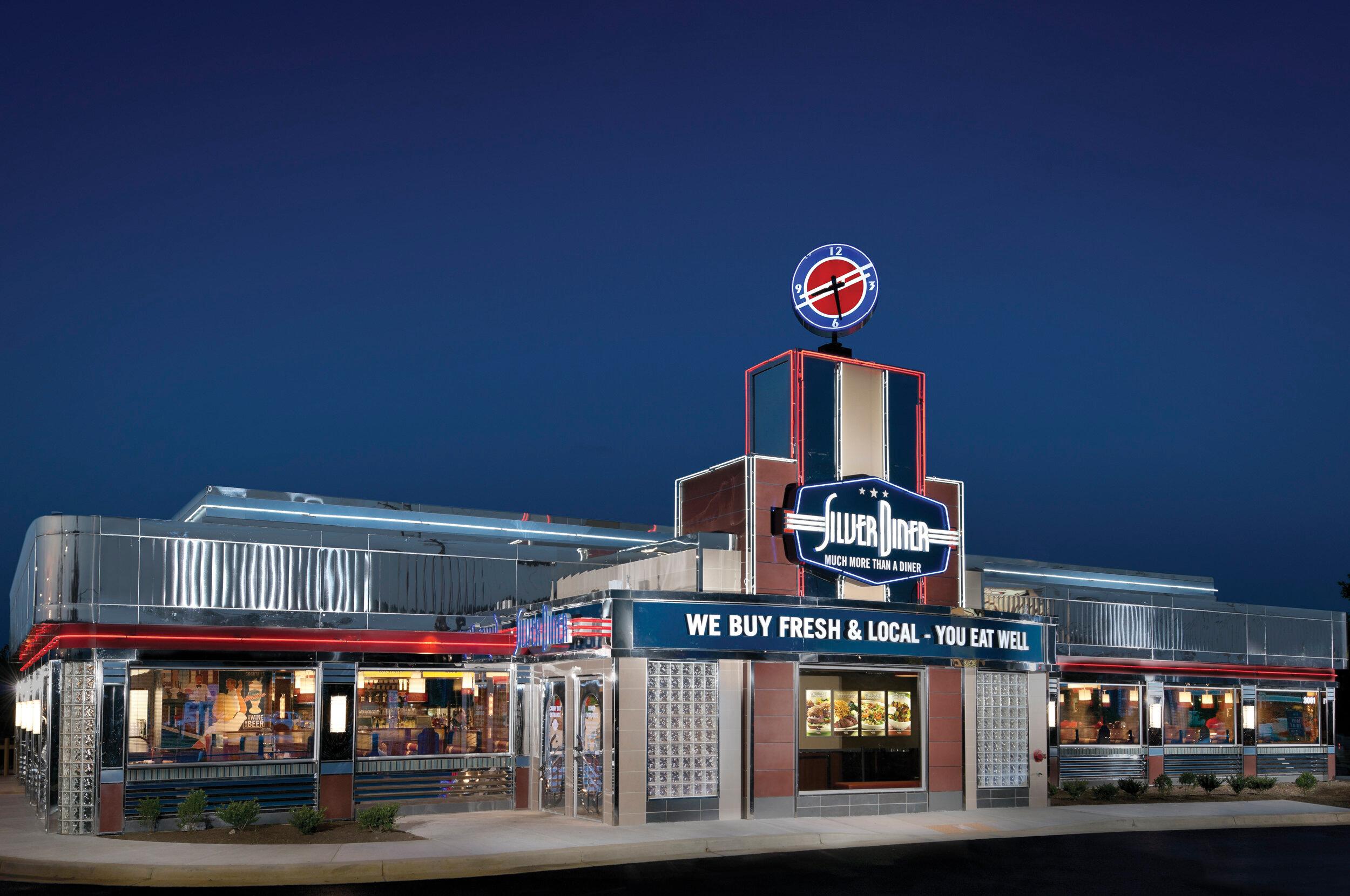Silver Diner exterior with shiny chrome design, neon lights, rooftop clock, and signage reading “Much More Than A Diner” and “We Buy Fresh & Local – You Eat Well.” Interior visible through windows with diners and staff in classic setting.