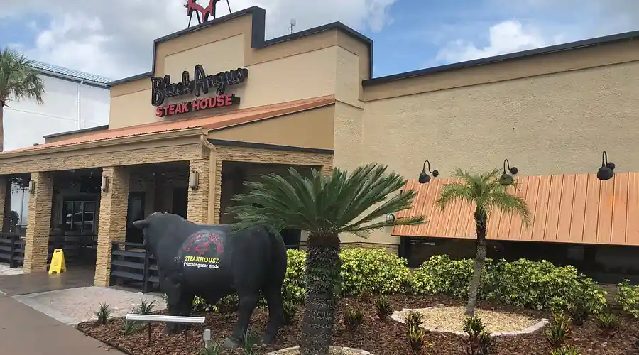 Exterior of Black Angus Steakhouse featuring beige facade, bold signage, a black cow statue, and lush landscaping with palm trees under a partly cloudy sky.