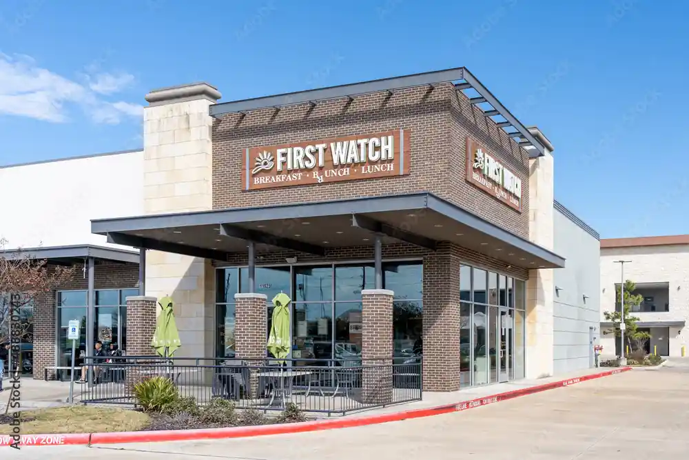 Daylight photo of First Watch restaurant exterior showing brick and light stone facade, signage reading “FIRST WATCH BREAKFAST • BRUNCH • LUNCH,” green umbrella-covered outdoor seating area, surrounding parking lot, and landscaping with small plants under partly cloudy skies.