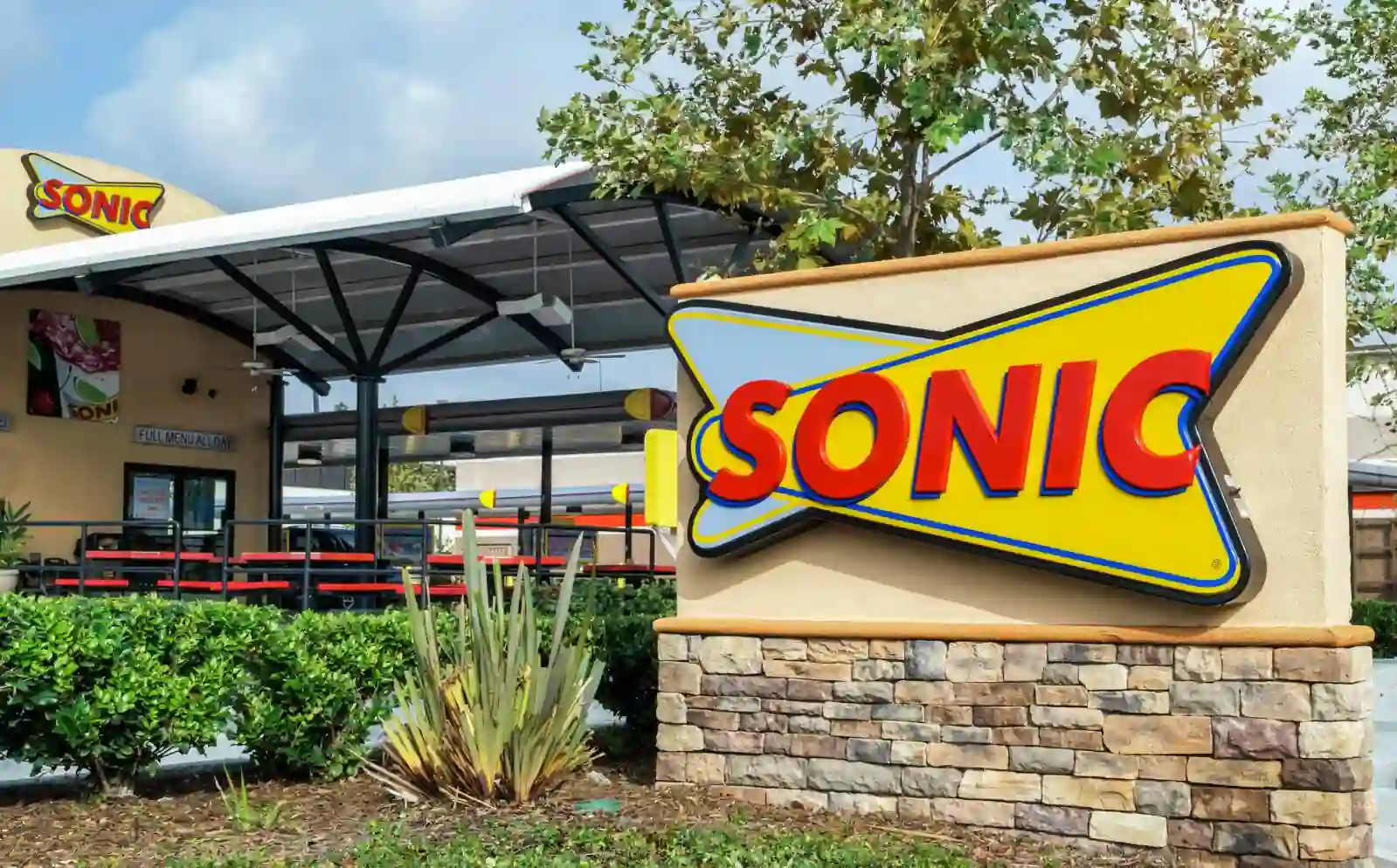 Colorful Sonic Drive-In exterior with a large logo sign, red benches under a canopy, and landscaped greenery.