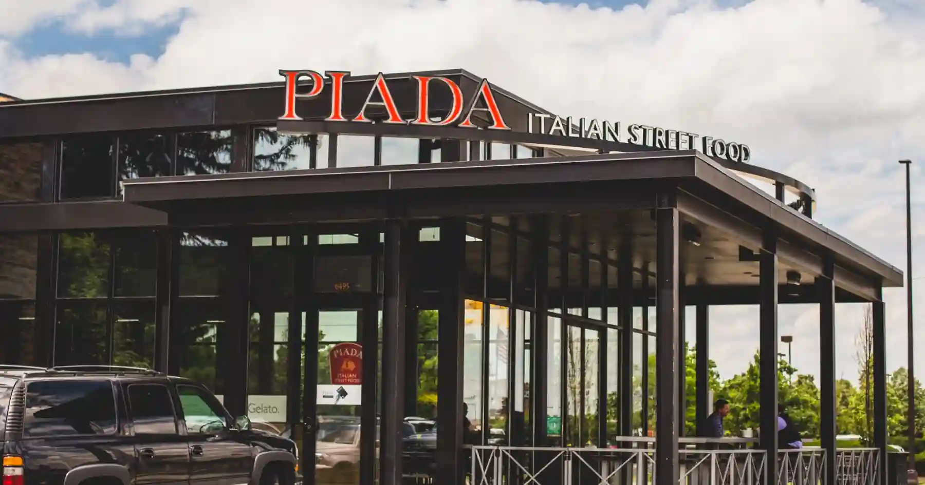 Exterior of Piada Italian Street Food restaurant with red signage, glass-panel facade, black metal frame, and outdoor seating area. Black SUV parked in front; entrance labeled with address number “6495.”