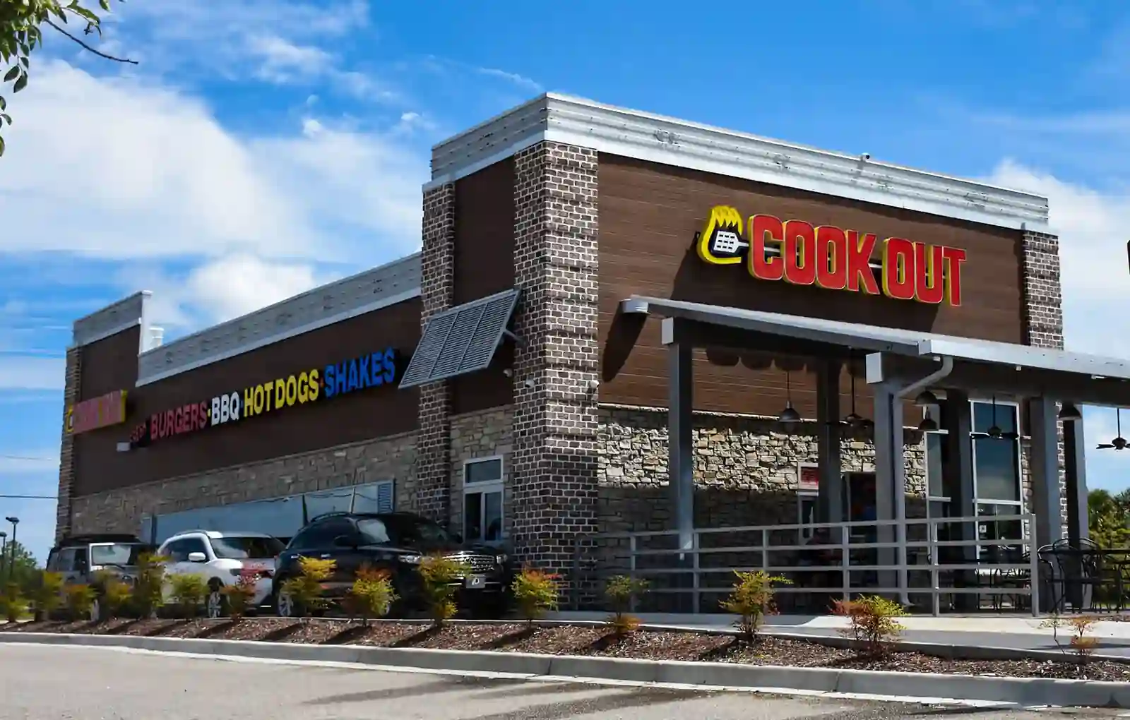 Exterior of Cook Out restaurant with red flame logo signage, brick and stone building features, smaller signs for burgers, BBQ, hot dogs, and shakes, cars parked in front, and landscaping with bushes under a mostly clear sky.