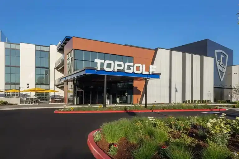 You Uploaded image Copilot The image shows the exterior of a Topgolf facility. The building has a modern design with large glass windows and a combination of white, brown, and gray panels. The entrance is marked by a large blue sign with the word "TOPGOLF" in white letters. To the left of the entrance, there is an outdoor seating area with yellow umbrellas. The landscaping in the foreground includes various plants and flowers, and the area is bordered by a red curb. The sky is clear and blue, indicating a sunny day.