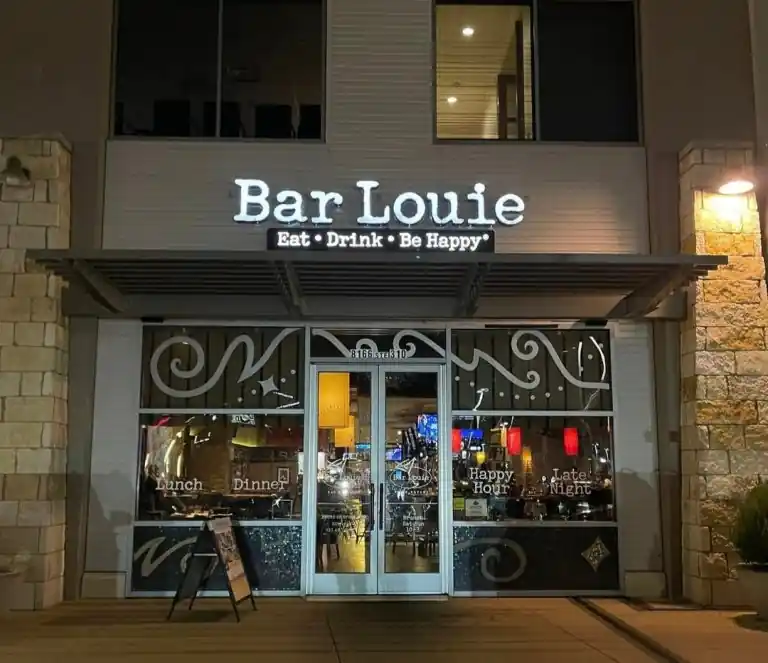 he image shows the exterior of a restaurant and bar named "Bar Louie." The sign above the entrance reads "Bar Louie" in large, illuminated letters, with the tagline "Eat • Drink • Be Happy" below it. The entrance features glass doors and windows with decorative swirls and text indicating offerings like "Lunch," "Dinner," "Happy Hour," and "Late Night." The address "8166" is visible above the door. Inside, the bar is dimly lit, with bar stools, tables, and hanging lights. The building has a modern design with stone and metal elements.