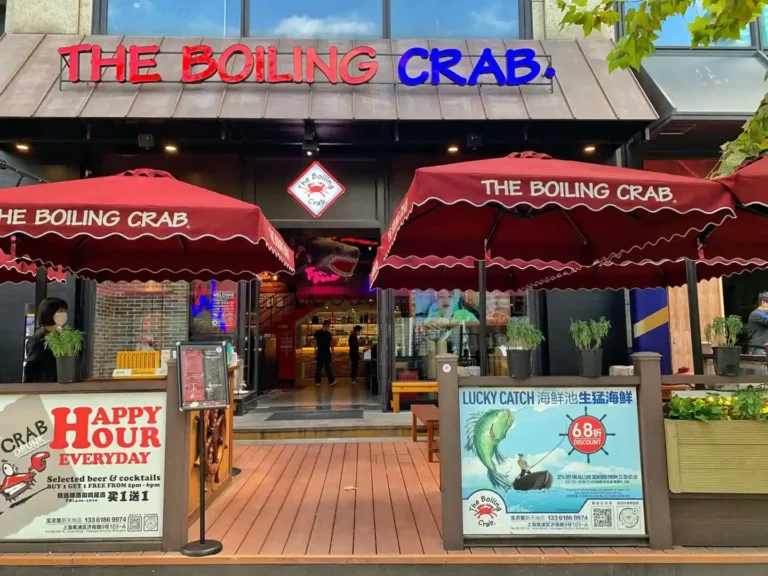 The image shows the exterior of a restaurant named "The Boiling Crab." The restaurant has a prominent sign with the name "THE BOILING CRAB" in red and blue letters. There are two large red umbrellas with the restaurant's name on them, providing shade for the outdoor seating area. The entrance to the restaurant is open, revealing the interior where a few people can be seen. There are two signs in front of the restaurant: The sign on the left reads: "HAPPY HOUR EVERYDAY Selected beer & cocktails BUY 1 GET 1 FREE FROM 2pm - 6pm" The sign on the right reads: "LUCKY CATCH 68折 DISCOUNT 20% OFF ON ALL LIVE SEAFOOD FROM 7:30-22:30 The Boiling Crab 133 6186 9974 上海市静安区南京西路1038号 近江宁路"