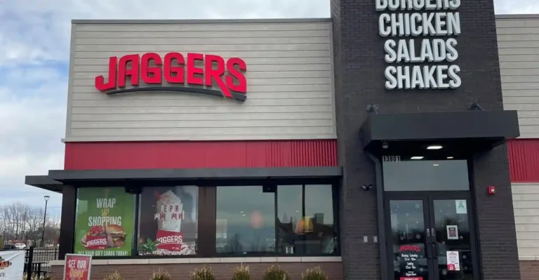 The image shows the exterior of a restaurant named "Jaggers." The restaurant has a sign with the name "JAGGERS" in large red letters on the left side of the building. On the right side, there is a vertical sign listing menu items: "BURGERS," "CHICKEN," "SALADS," and "SHAKES." Below this sign is the entrance door with the address number "13801" above it. The door has the restaurant's hours listed: "Monday - Saturday 11am - 9pm, Sunday 11am - 8pm." There are also promotional posters in the windows advertising gift cards and a "PEPPERMINT SHAKE." The building has a combination of light grey and dark grey exterior walls with red accents.