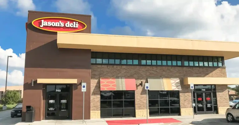 The image shows the exterior of a Jason's Deli restaurant. The building has a modern design with a combination of brick and brown paneling. The sign for Jason's Deli is prominently displayed at the top left corner of the building. The entrance doors have the number "742" above them, and there are two handicap parking signs in front of the building. The windows have decorative awnings with geometric patterns. The sky is clear with some clouds, indicating a sunny day.