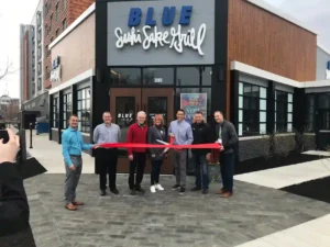 Ribbon-cutting ceremony in front of BLUE Sushi Sake Grill, with six individuals holding a red ribbon and a large pair of scissors.