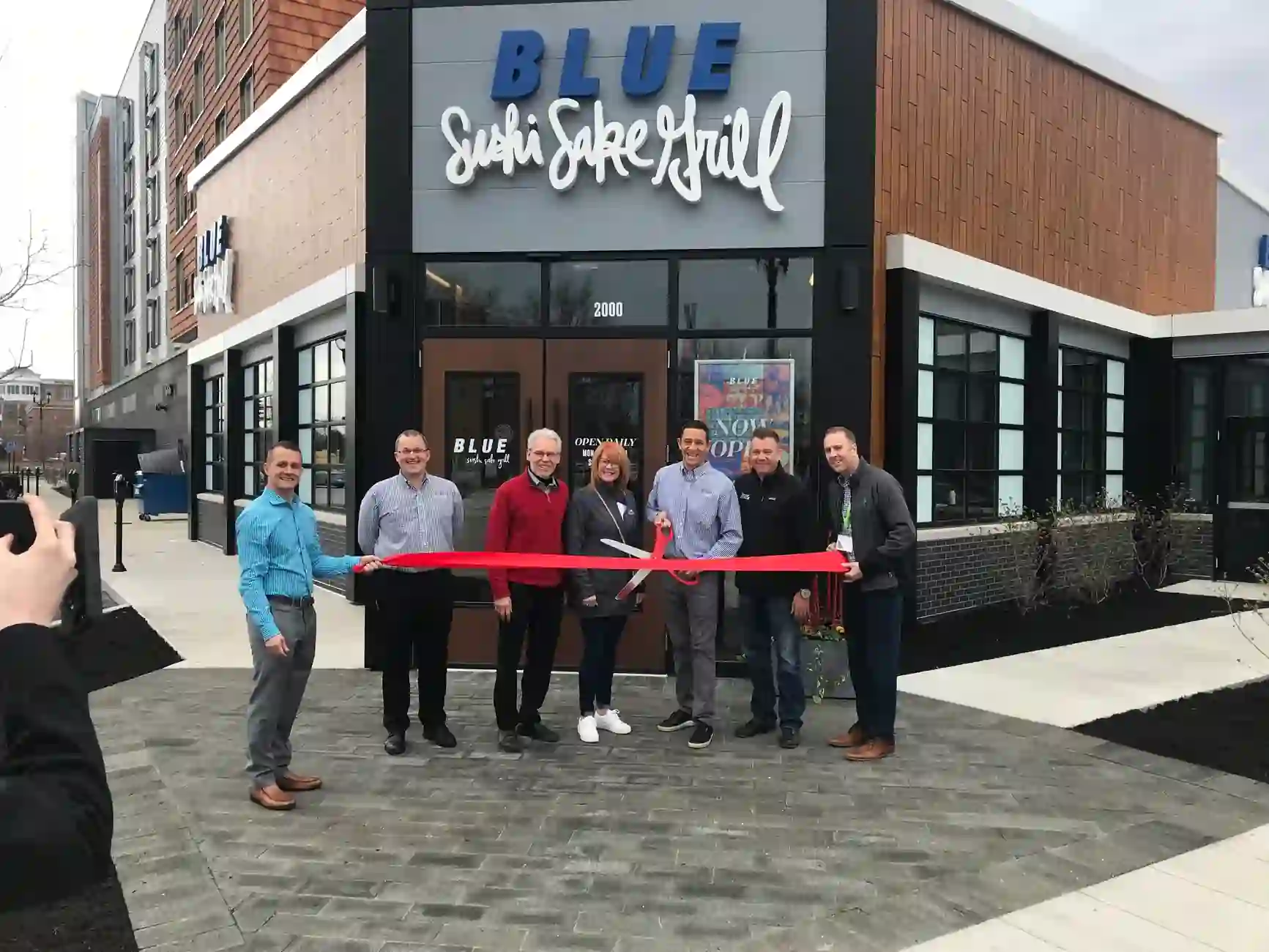 Ribbon-cutting ceremony in front of BLUE Sushi Sake Grill, with six individuals holding a red ribbon and a large pair of scissors.