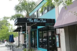 Exterior of The Blue Fish restaurant featuring a teal-painted building, black awning with light blue cursive text, and outdoor seating under blue umbrellas.