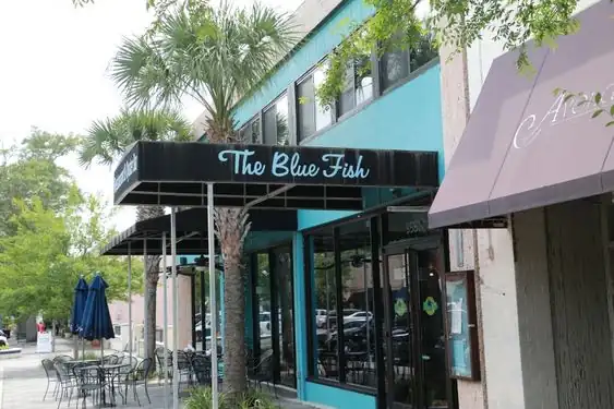 Exterior of The Blue Fish restaurant featuring a teal-painted building, black awning with light blue cursive text, and outdoor seating under blue umbrellas.