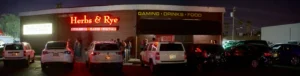 Exterior view of Herbs & Rye steakhouse at night with a crowd and parked cars in front.