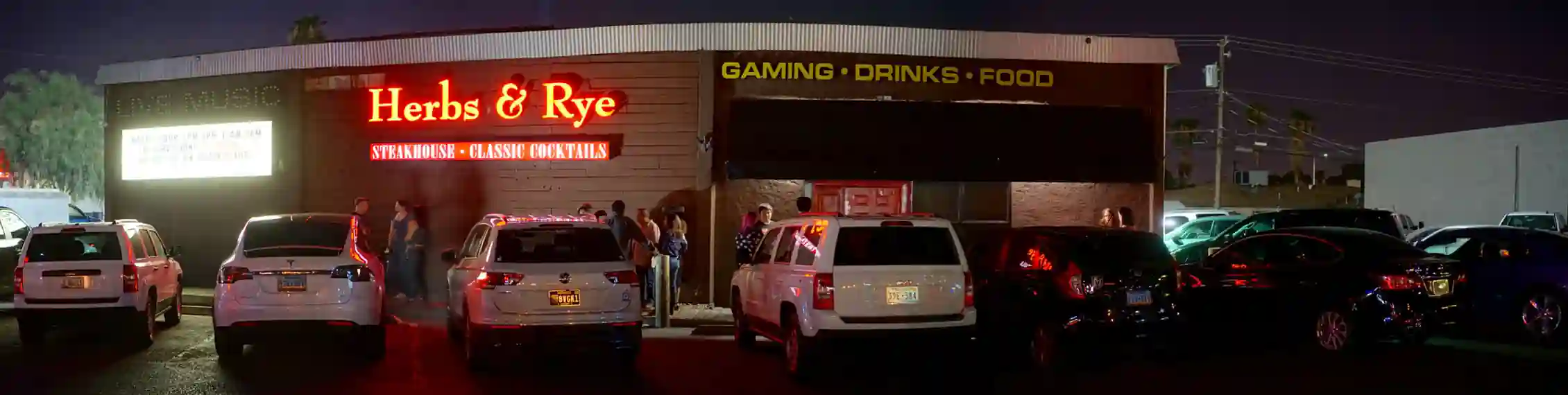 Exterior view of Herbs & Rye steakhouse at night with a crowd and parked cars in front.