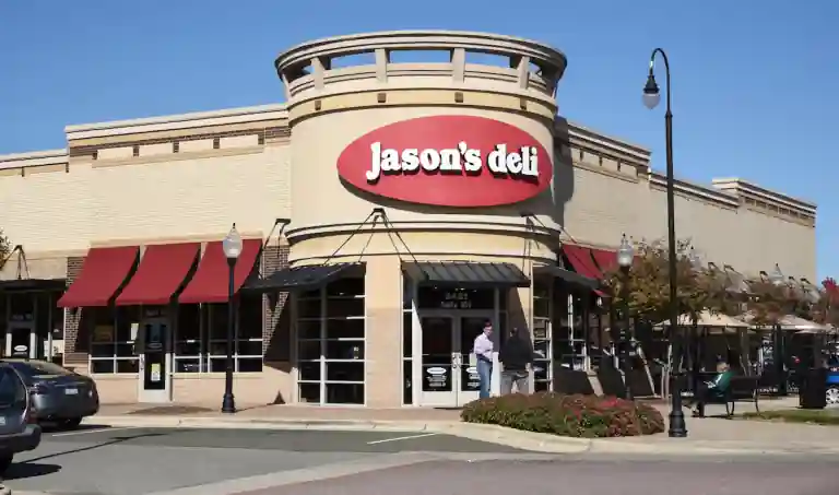 Front view of Jason's Deli restaurant featuring a beige brick facade with a large red oval logo reading 'Jason's Deli' in bold white letters. The entrance has a round architectural tower design, black metal awning, and glass doors. Red window awnings line the side windows. Two people are seen near the entrance, with cars parked in front. Decorative lamp posts, trees, and bushes enhance the outdoor atmosphere under a clear blue sky.
