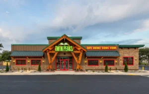 Twin Peaks restaurant exterior featuring a lodge-style design with a neon green sign, stone accents, wooden beams, and a green metal roof.