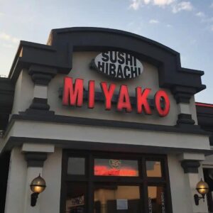 The exterior of Miyako Sushi & Hibachi restaurant with illuminated red signage, decorative columns, and wall-mounted lamps. The entrance features a neon “Restaurant” sign, with the sky visible in the background.