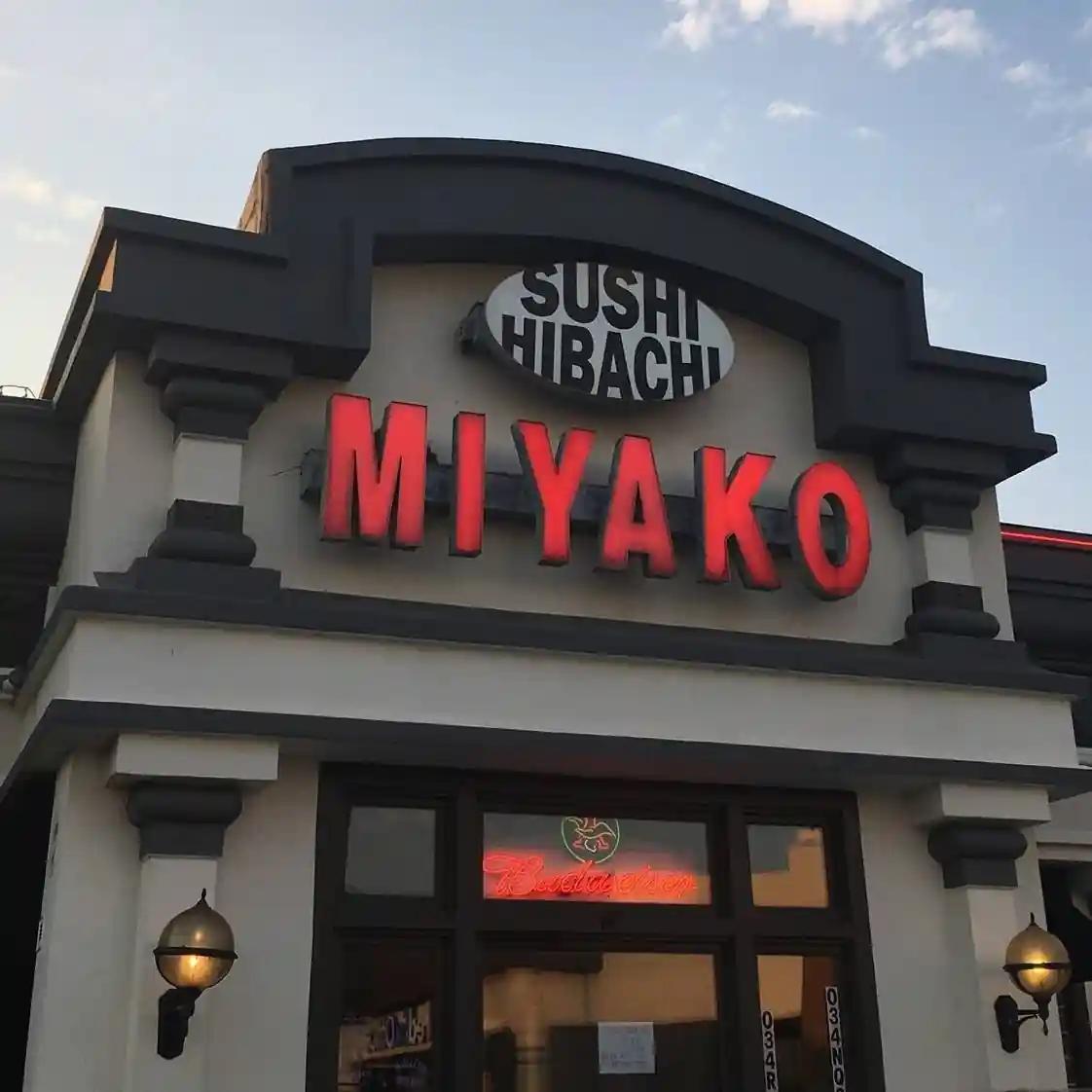 The exterior of Miyako Sushi & Hibachi restaurant with illuminated red signage, decorative columns, and wall-mounted lamps. The entrance features a neon “Restaurant” sign, with the sky visible in the background.