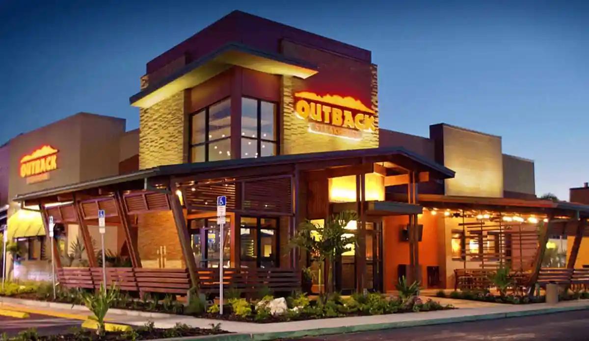 Exterior of an Outback Steakhouse restaurant with modern stone and wood design, illuminated signage, and evening lighting.