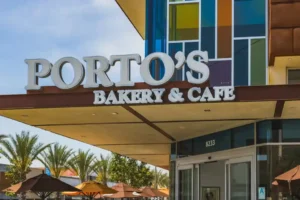 Exterior of Porto's Bakery & Cafe with a bold sign, glass entrance, colorful facade, and outdoor seating area with umbrellas and palm trees.
