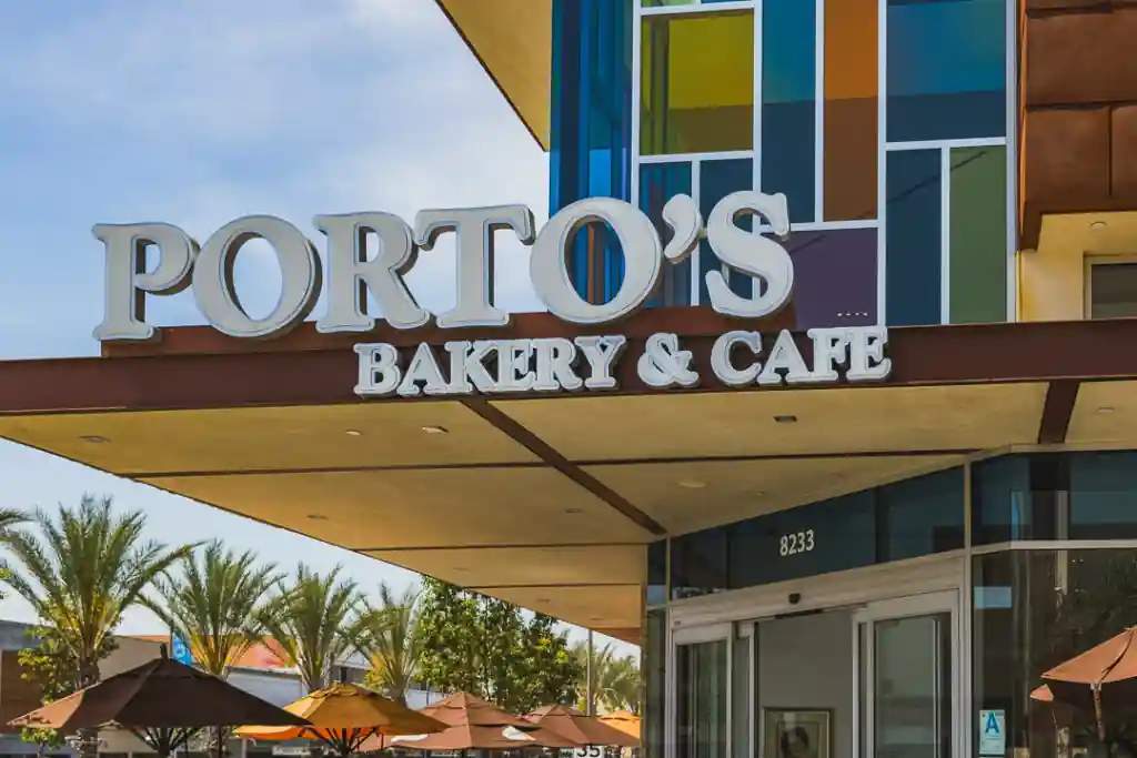 Exterior of Porto's Bakery & Cafe with a bold sign, glass entrance, colorful facade, and outdoor seating area with umbrellas and palm trees.