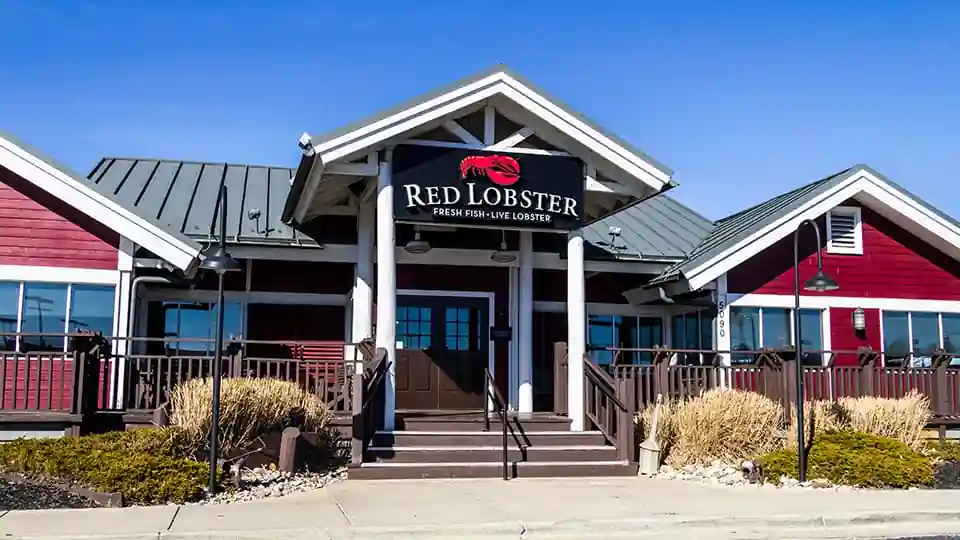 The exterior of a Red Lobster restaurant with a red and white color scheme, a gray roof, and a bold sign above the entrance featuring a red lobster graphic alongside the words "RED LOBSTER." Below, the sign reads "FRESH FISH - LIVE LOBSTER." The entrance has double doors and is flanked by windows. A small porch with railings and landscaped bushes completes the scene.