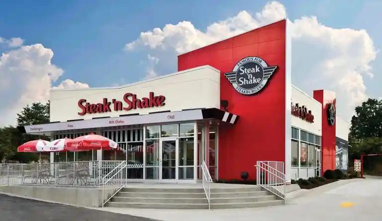 Exterior of a Steak 'n Shake restaurant featuring a modern red and white design, large signage, and outdoor seating with Coca-Cola branded umbrellas.