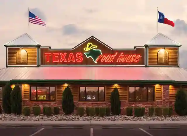 The image displays the exterior of a Texas Roadhouse restaurant, featuring a rustic wooden facade and a metal roof. The restaurant's name is illuminated in red and cursive neon, alongside a green outline of the state of Texas and a yellow cowboy hat. Two flags—the American flag and the Texas state flag—are mounted on top.