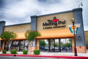 The exterior of The Melting Pot, a fondue restaurant, with a beige and gray facade, orange awning, and glass doors. Two small trees with red flowers and a lamp post with a decorative banner are in front.