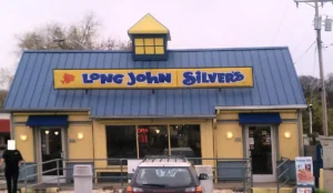 The exterior of a Long John Silver's restaurant with a blue roof, yellow signage, and two entrances with blue awnings. A car is parked in front, and a promotional sign advertises a meal deal.