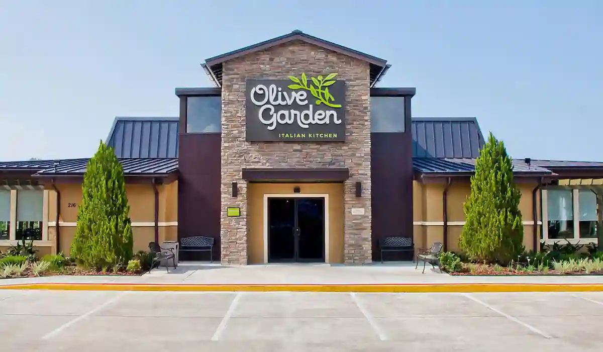 The image features the exterior of an Olive Garden Italian Kitchen restaurant. The building showcases a rustic stone facade, with a large sign displaying the restaurant’s name and olive branch logo. In front of the entrance, there are two benches and some neatly arranged greenery.