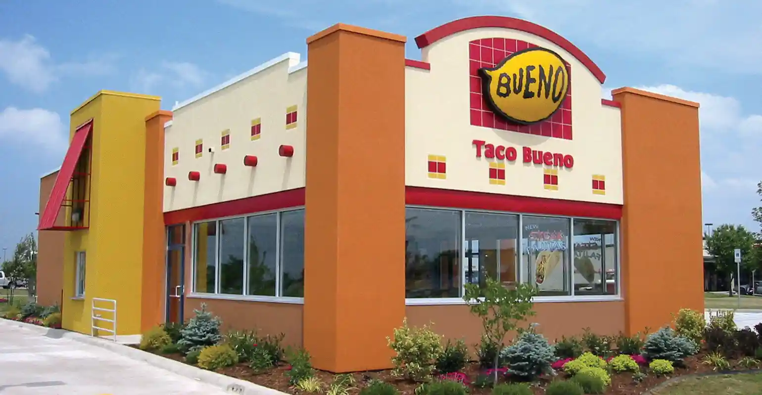 "Exterior view of a Taco Bueno restaurant featuring a vibrant yellow, orange, and red color scheme. The building has a bold design with large orange columns, red accents, and a distinctive yellow oval sign with the word 'BUENO' in bold black letters. Large glass windows display promotional posters. Landscaped greenery and flowers surround the restaurant, with a bright blue sky overhead.