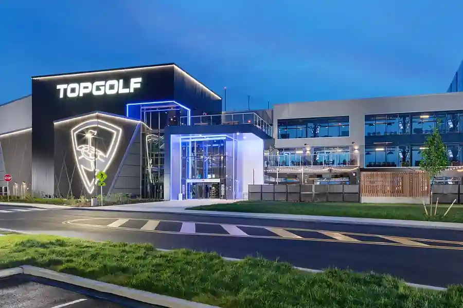 Topgolf building at twilight featuring illuminated signage, large glass entrance, multi-story design, and golf-themed logo; visible stop sign and pedestrian crossing in foreground.