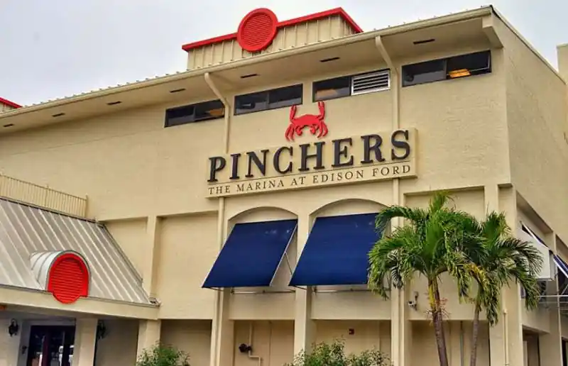 Exterior of Pinchers restaurant featuring red crab logo, beige walls, blue awnings, palm tree, and signage reading "The Marina at Edison Ford" — representing a waterfront dining venue.