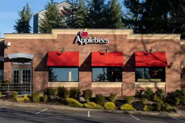 Front exterior of Applebee’s restaurant with red brick facade, red apple logo signage, pink window awnings, and green benches beneath a cloudy sky.