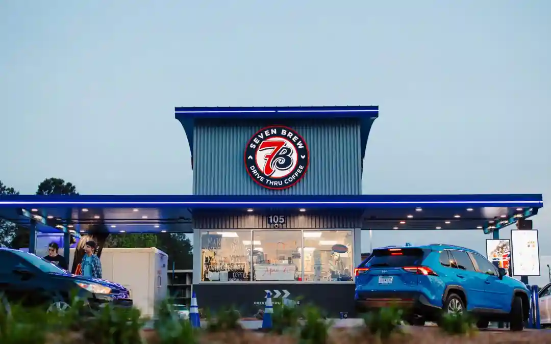 Nighttime exterior of Seven Brew Drive Thru Coffee with illuminated circular logo, modern blue-accented structure, cars in drive-thru, and “105” address over window. Handwritten menu visible behind glass with customers at service window.