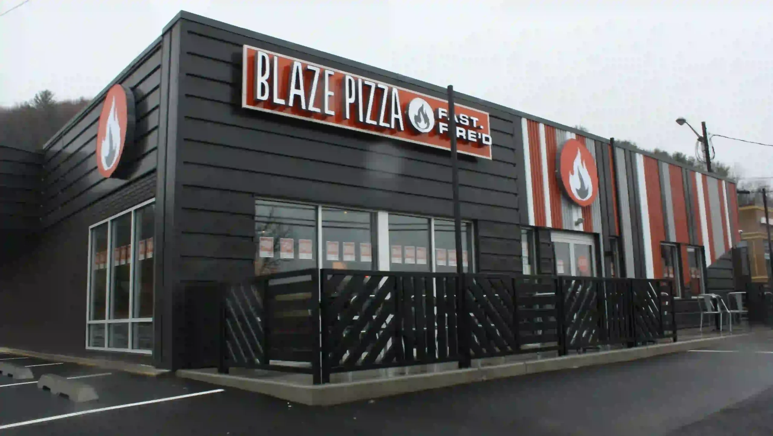 Blaze Pizza restaurant exterior with modern black paneling, red and gray vertical accent stripes, large Blaze Pizza flame logo, and “FAST FIRE’D” signage above tall glass windows and patio railing.