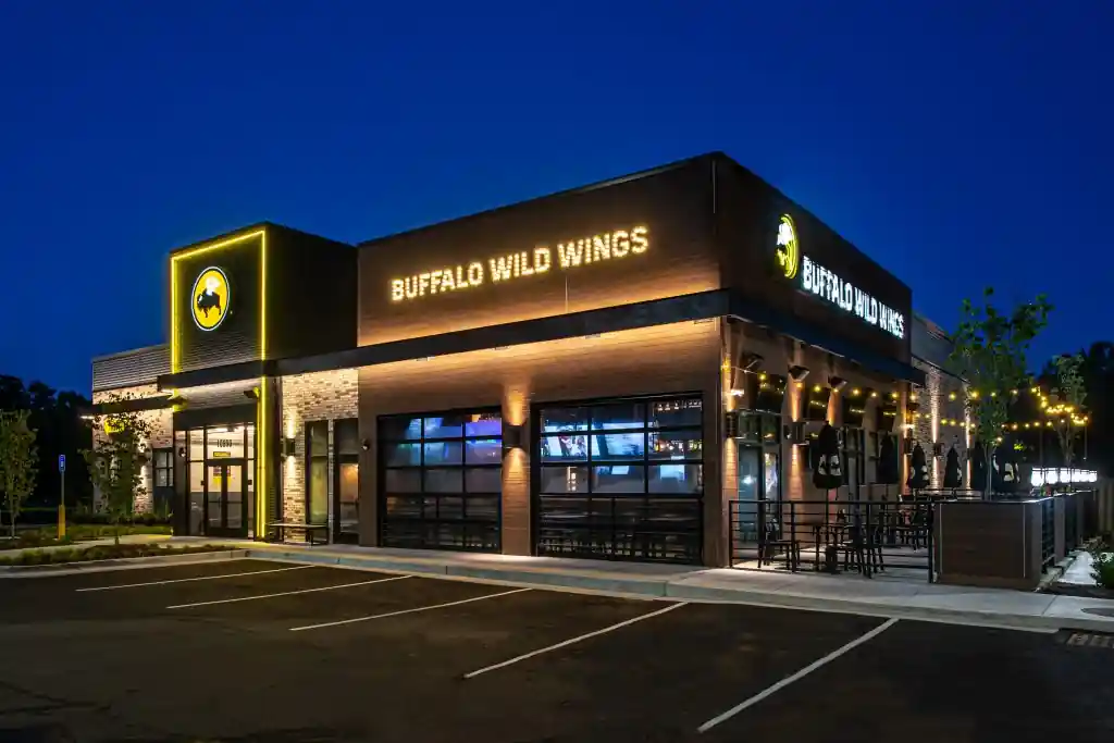 Nighttime view of Buffalo Wild Wings restaurant with illuminated signage, yellow buffalo logo, outdoor seating area, and an empty parking lot under a dark sky.